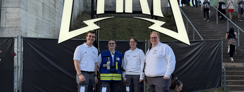 4 ehrenamtliche EInsatzkräfte des BRKs stehen vor dem Rock im Park Logo-Schild.